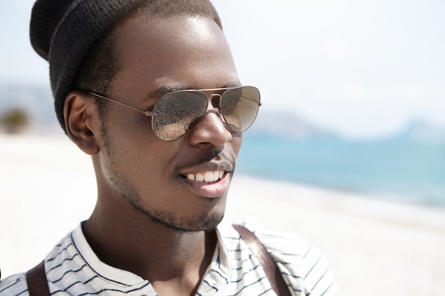 Free photo close up shot of happy young african american backpacker in mirrored lens sunglasses relaxing on beach on sunny day