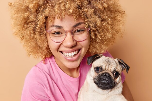 Close up shot of happy young woman with curly hair smiles gladfully enjoys company of pet poses with pug dog dressed casually isolated over beige background People and domestic animals concept