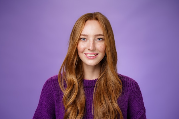 Close-up shot of hopeful and optimistic happy young redhead 20s girl with freckles and long hair smiling joyfully with faith in eyes and prominent look posing against purple background.