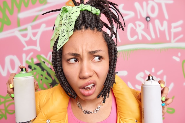 Close up shot of indignant dissatisfied Afro American teenage girl purses lips concentrated aside has dreadlock hairstyle holds aerosol bottles makes creative paintingson street wall