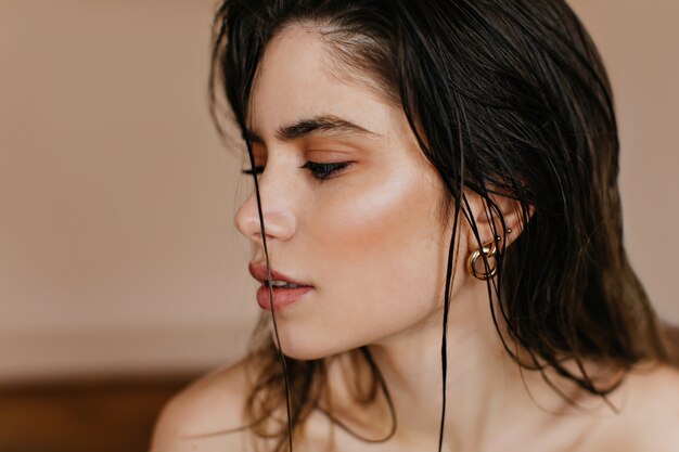 Close-up shot of inspired european woman with golden earrings. Indoor photo of interested brunette lady posing on brown wall.