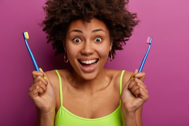 Close up shot of joyful Afro American woman holds two tooth brushes