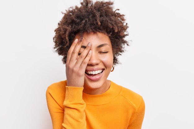 Close up shot of joyful carefree young woman with curly Afro hair smiles toothily keeps eyes closed makes face palm wears orange jumper expresses happiness isolated over white  wall