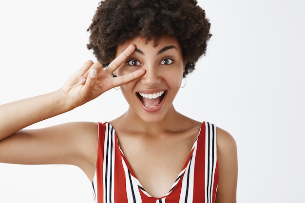 Free photo close-up shot of joyful optimistic and happy disco girl with afro hairstyle in striped blouse showing victory or peace sign over eye and smiling broadly feeling happy and amused