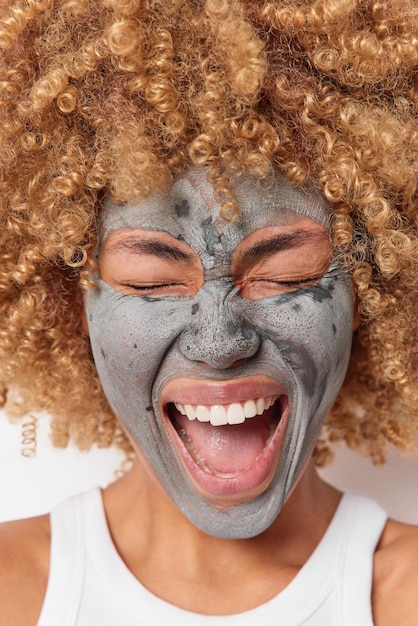 Close up shot of joyful woman exclaims from happiness has overjoyed expression applies clay mask for reducing fine lines poses indoor undergoes skin care procedures. Beauty treatments concept
