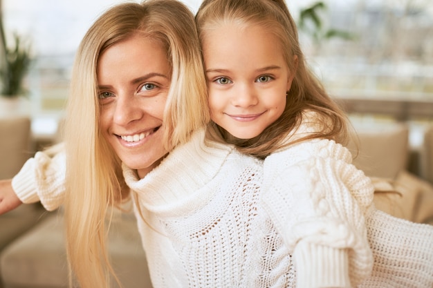 Close up shot of joyful young blonde female in white sweater giving back ride yo her adorable baby daughter spending winter December day at home, laughing, bonding and entertaining themselves