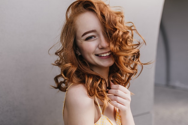 Close-up shot of laughing jocund lady posing on gray wall. Photo of winsome ginger girl with elegant curly hairstyle.