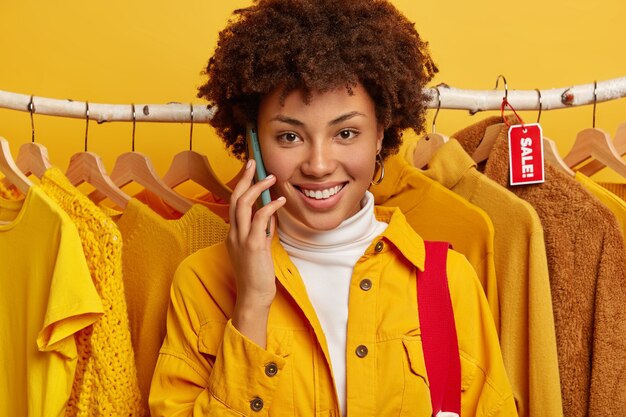 Close up shot of lovely curly haired woman makes phone call, smiles broadly, dressed in yellow jacket