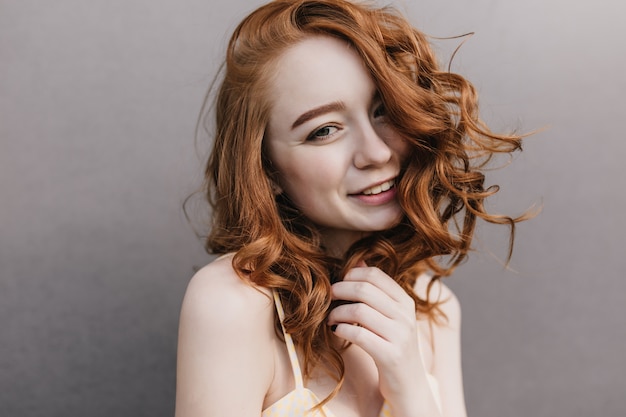 Close-up shot of lovely ginger woman smiling. Portrait of red-haired curly girl isolated on gray wall.