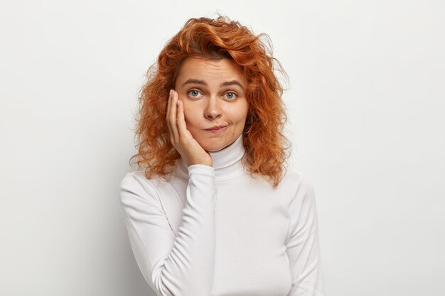 Close up shot of lovely woman with red curly hair, purses lips and looks with doubtful expression, touches cheek, poses against white wall, wears poloneck jumper. Human facial expressions