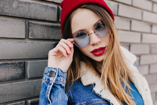 Close-up shot of magnificent woman with light-brown hair touching her blue glasses. Outdoor photo of charming girl in denim jacket standing on urban wall in cold morning.