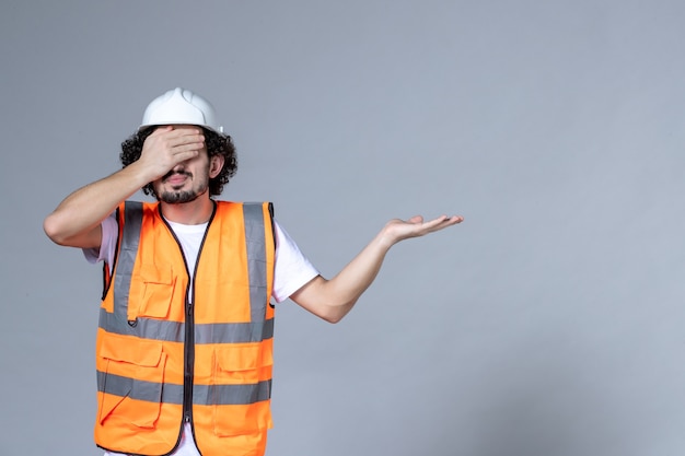 Foto gratuita immagine ravvicinata del costruttore maschio in giubbotto di avvertimento che indossa il casco di sicurezza e indica qualcosa sul lato sinistro chiudendo gli occhi sul muro grigio dell'onda