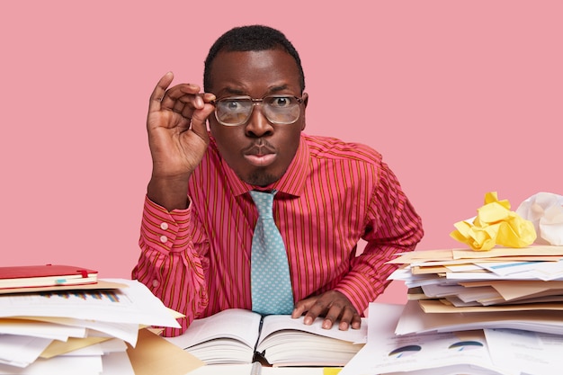 Free photo close up shot of male wonk looks scrupulously, keeps hand on rim of spectacles, dressed in formalwear, sits at desktop, reads literature