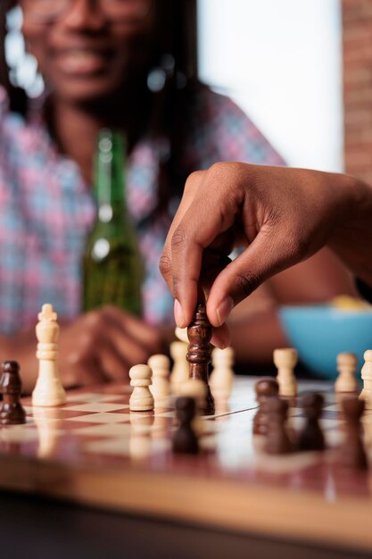 Close up shot of man moving chess piece on chessboard while sitting at table. Smart person playing strategy boardgame at home in living room while having snacks and beverages.