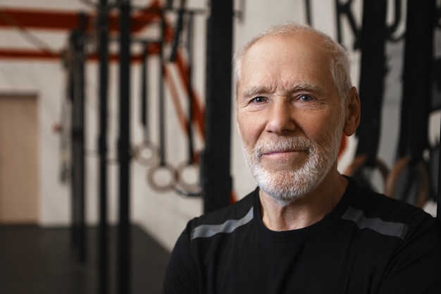 Close up shot of mature seventy year old Caucasian man with wrinkles, blue eyes and thick beard wearing stylish black t-shirt while exercising in gym alone, looking at camera