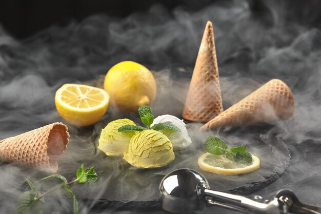 Close-up shot of a natural appetizing creamy and lemon ice cream decorated with mint and served in smoke on a dark stone slate over a black background. Metal scoop is laying nearby.