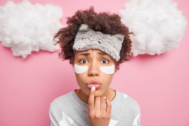 Close up shot of nervous Afro American woman keeps finger on folded lips dressed in nightwear wears sleepmask on forehead isolated over pink wall white clouds above