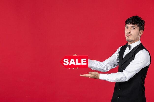 Close up shot of nervous guy waiter in a uniform with butterfly on neck showing sale icon pointing something on the right side on isolated red background