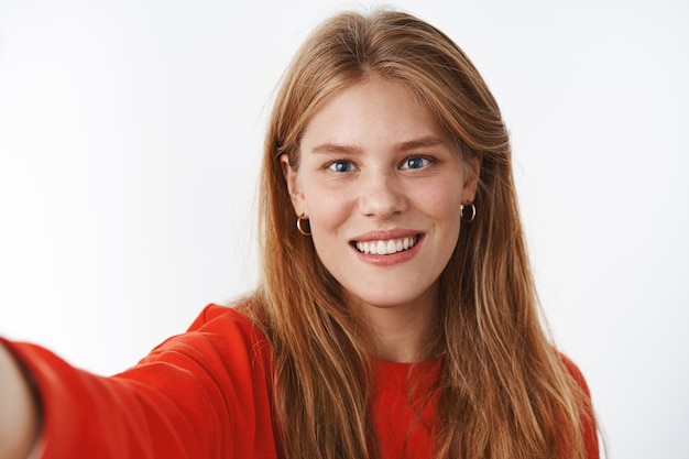 Close-up shot of nice tender young fair-haired woman with freckles, chubby cheeks and blue eyes pulling hand forward, holding camera and smiling as if taking selfie, posing over gray wall