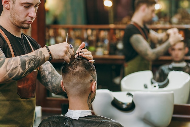 理髪店​で​流行​の​散髪​を​している​男性​の​クローズアップ​ショット​。​クライアント​に​サービス​を​提供する​入れ墨​の​男性​の​ヘア​スタイリスト​。