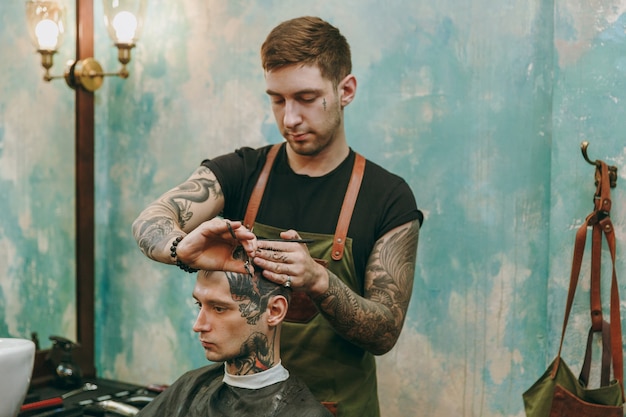 理髪店​で​流行​の​散髪​を​している​男性​の​クローズアップ​ショット​。​クライアント​に​サービス​を​提供する​入れ墨​の​男性​の​ヘア​スタイリスト​。