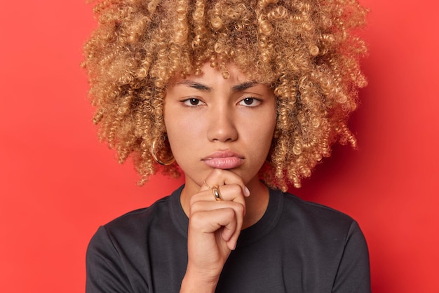 Close up shot of offended young woman keeps hand on chin purses lips looks sadly at camera looks directly at camera with sulking expression wears black t shirt isolated over vivid red background.