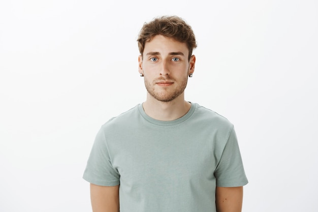 Close-up shot of ordinary fair-haired caucasian male with bristle in earrings, staring carefree being indifferent and calm