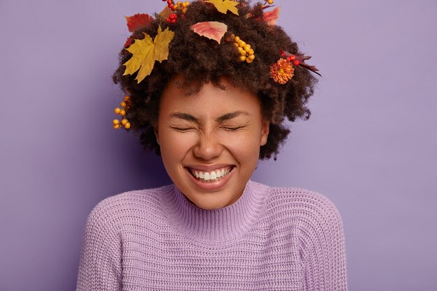 Close up shot of overjoyed curly woman has fun indoor,  being entertained, closes eyes with satisfaction and joy, shows white teeth, autumnal foliage in head