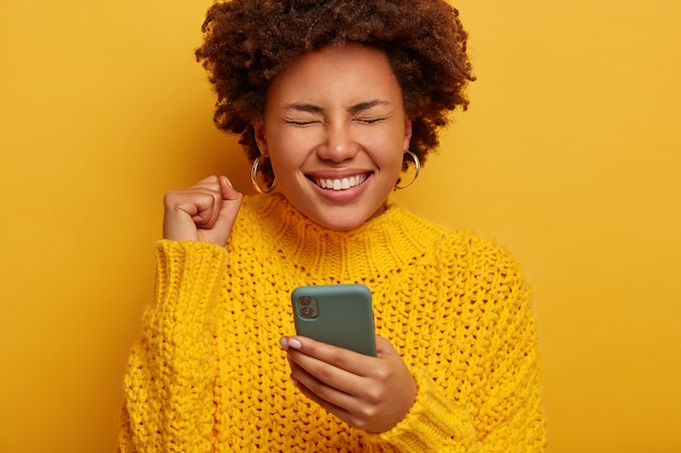 Close up shot of overjoyed curly woman keeps fist clenched, happy to get money reward, gets notification on cellphone, wears yellow knitted sweater