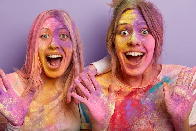 Close up shot of overjoyed emotional two women open mouth, exclaim with joy, have colored party, smeared each other with colorful powder, amazed by seeing something incredible on Indian color fest