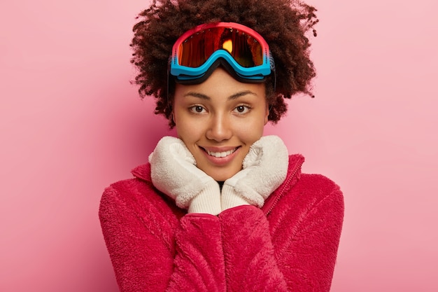 スノーボードマスクで見栄えの良い若い女性のクローズアップショット、あごの下で両手を保持し、暖かい白い手袋を着用し、カメラを直接見ています。