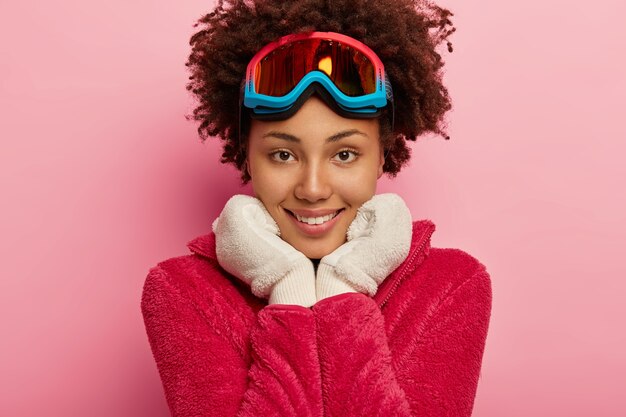 Close up shot of pleasant looking young woman with snowboard mask, holds both hands under chin, wears warm white gloves, looks directly at camera.