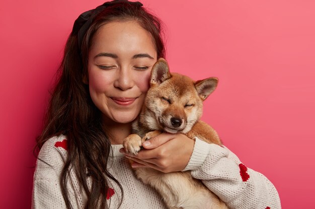 Close up shot of pleased Asian woman carries pedigree dog closely near face, closes eyes from pleasure, embraces animal with love