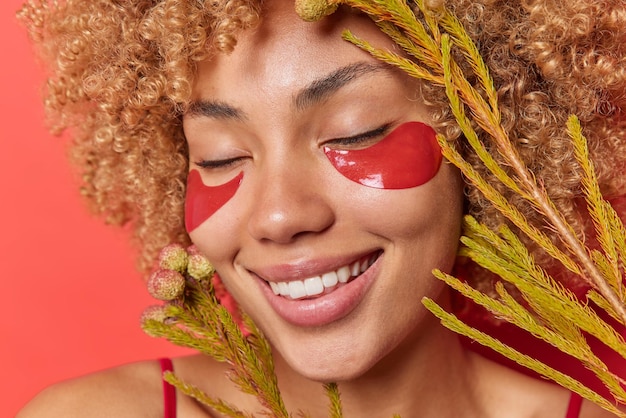 Крупный план довольной молодой женщины со здоровой кожей держит глаза закрытыми, применяет красные гидрогелевые пластыри для ухода и увлажнения, держит экзотическое растение возле лица, позирует в помещении Концепция красоты