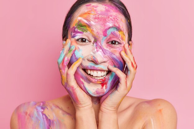 Close up shot of positive Asian woman smeared with colorful gouache paints keeps hands on cheeks smiles happily