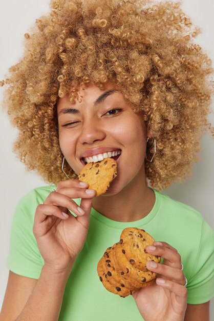Close up shot of positive curly haired young woman eats delicious appetizing cookies with chocolate winks eye dressed in casual green t shirt has sweet tooth addiction to sugar poses indoor