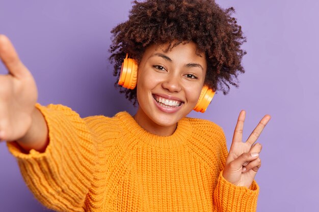 Close up shot of positive dark skinned ethnic woman makes victroy gesture smiles broadly and takes selfie listens audio track in wireless headphones 