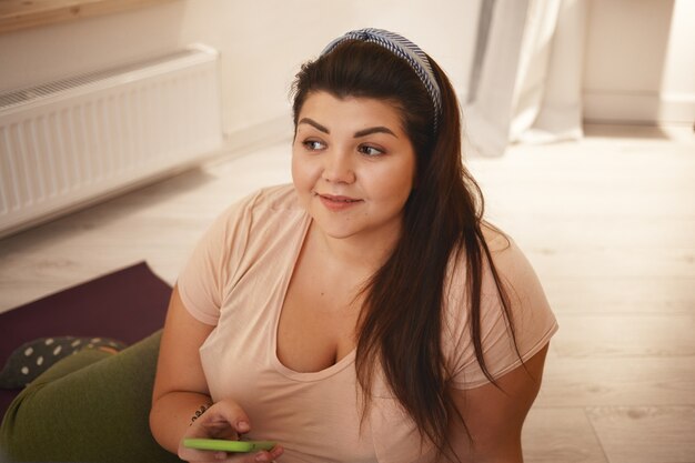Close up shot of positive young obese female with chubby cheeks and curvy body typing text message via online messenger using mobile phone after yoga practice, sitting on floor and smiling