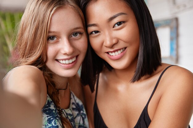 Close up shot of pretty delighted smiling woman being in high spirit, stand close to each other, smile joyfully, have friendly relationship.