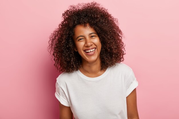 Close up shot of pretty teenage girl with dark skin, curly Afro hair, grins , has white teeth, laughs sincerely at good joke, has fun with close friend, wears everyday white t shirt