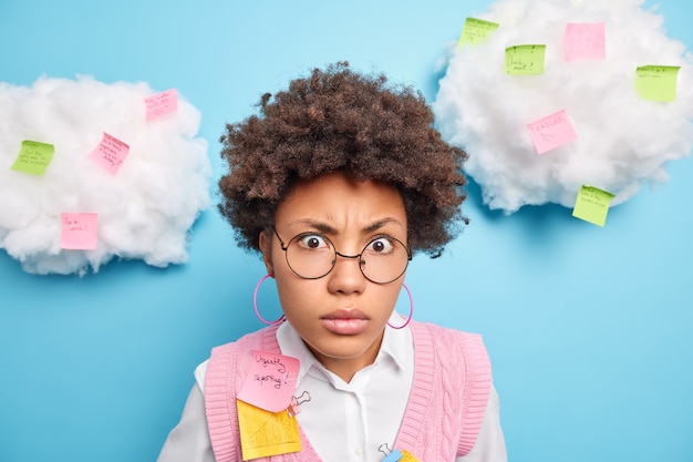 Close up shot of puzzled Afro American student looks shocked at camera wears round spectacles makes notes on stickers has different tasks to do isolated over blue wall