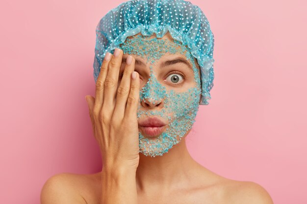 Close up shot of puzzled woman with blue scrub on face, covers one eye with hand, tries to hide herself, has stupefied expression, wears protective bathcap, wants to look younger, stands shirtless