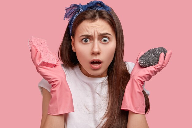 Close up shot of puzzled young woman has surprised facial expression, carries sponge, does household duties, amazed by amount of work