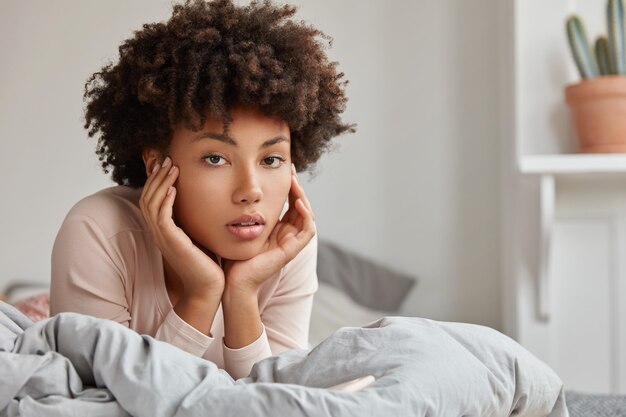 Снимок крупным планом расслабленной темнокожей женщины, которая лежит в постели, отдыхает в выходные или в выходной, смотрит прямо с задумчивым выражением лица