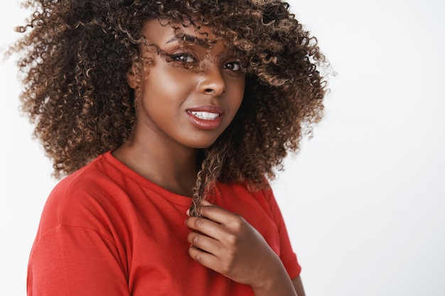 Close-up shot of romantic and sensual gorgeous dark-skinned woman with pure clean skin smiling tender touching curl of hair and waving haircut gazing soft and caring at front over white wall Free Photo