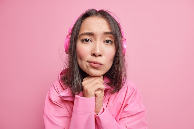 Close up shot of sad disappointed millennial girl keeps hands under chin looks with melancholic expression listens lyrics songs via headphones dressed in jacket isolated over pink wall