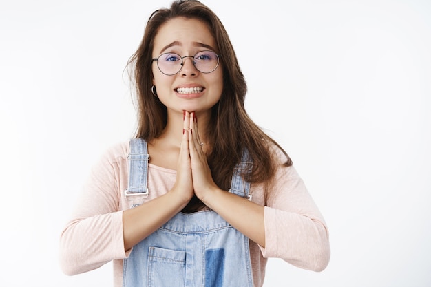 Close-up shot of sad and hopeful attractive woman in glasses standing in begging pose holding hands in pray expressing suppolication as wanting forgiveness, apologizing over gray wall.