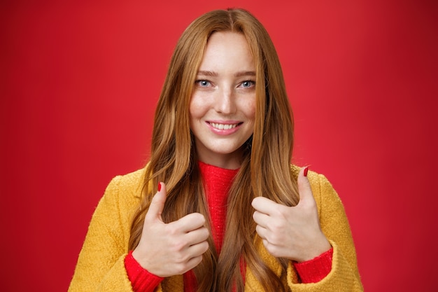 Free photo close-up shot of satisfied happy young 20s woman in yellow coat showing thumbs up gesture in like and approval giving positive feedback enjoying nice event standing over red background, smiling cute.