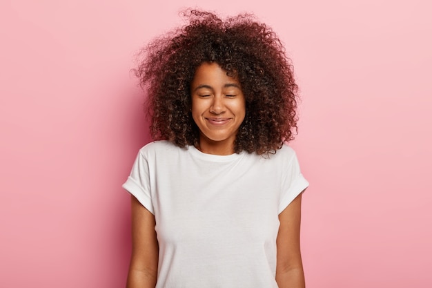 Close up shot of satisfied lovely teenager with bushy curly hair, has eyes shut, pleasant smile, awaits for surprise with great happiness, enjoys awesome time during weekend, wears white t shirt