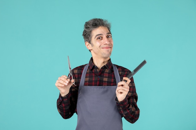 Close up shot of satisfied smilling young hairdesser wearing gray apron and holding scissor comb on pastel blue color surface